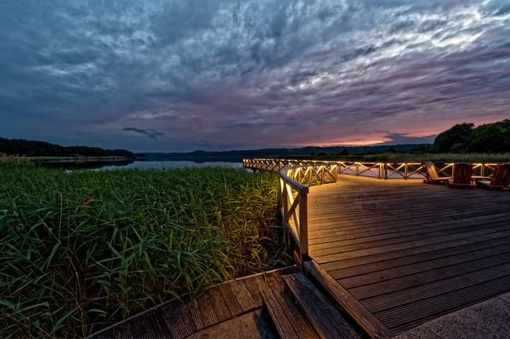 Holzsteg mit beleuchteten Handläufen, der in der Abenddämmerung unter bewölktem Himmel zu einem See führt.