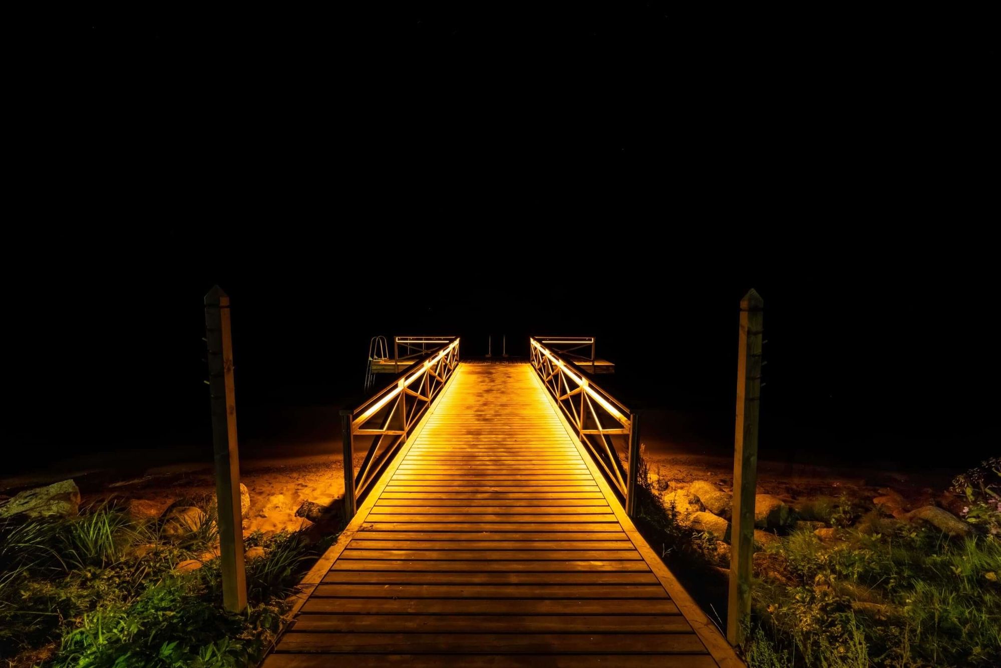 Beleuchteter Holzsteg bei Nacht, der in die Dunkelheit führt.