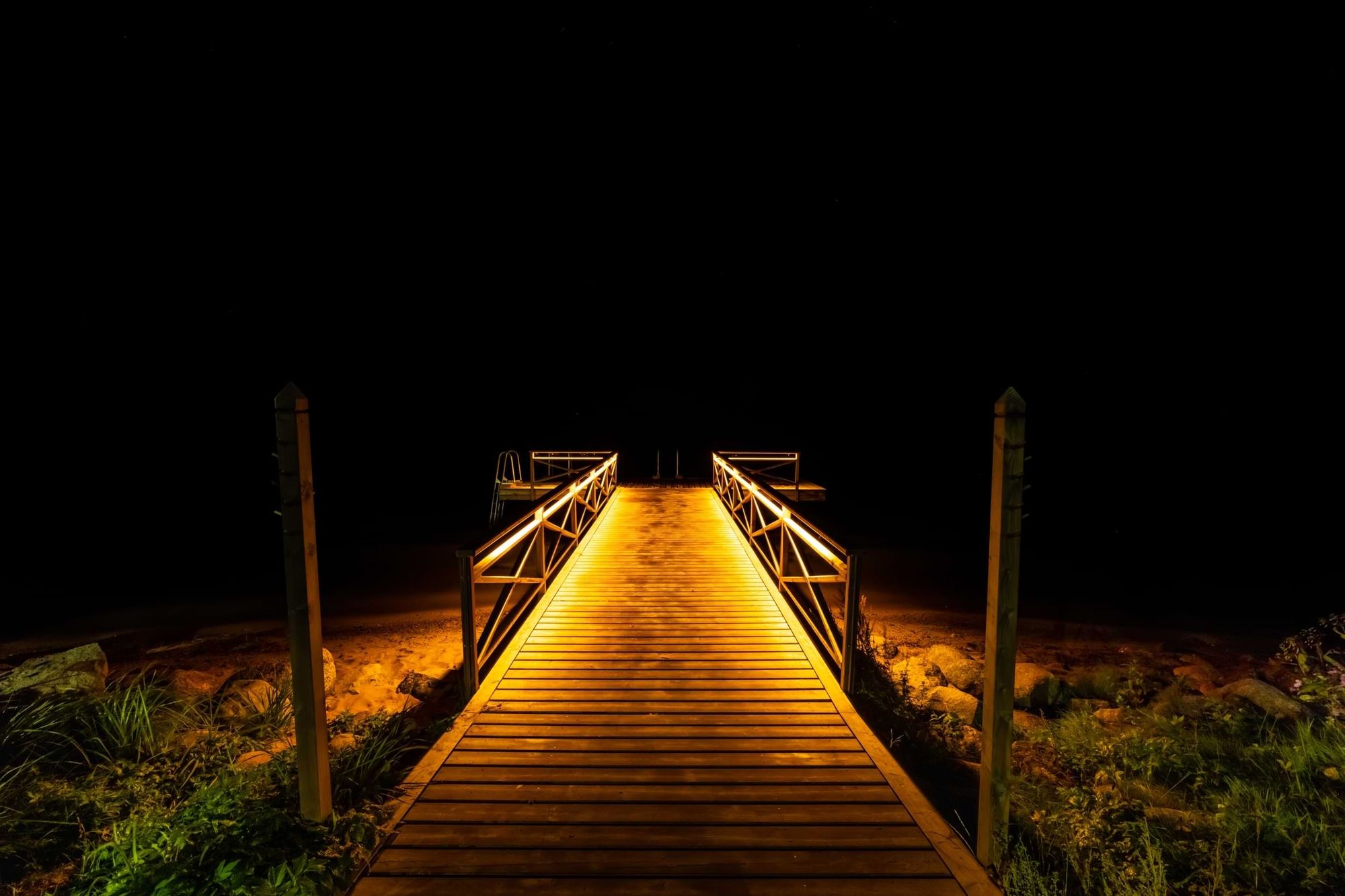 Beleuchteter Holzsteg bei Nacht, der in die Dunkelheit führt.