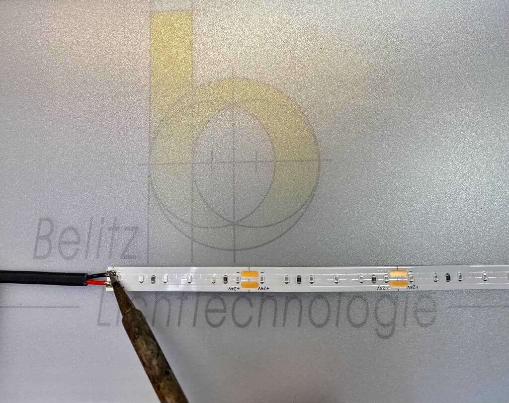 Ein Lineal, das mit einem Lötkolben auf einer grauen Metalloberfläche mit Text ein Band mit LED-Leuchten misst.